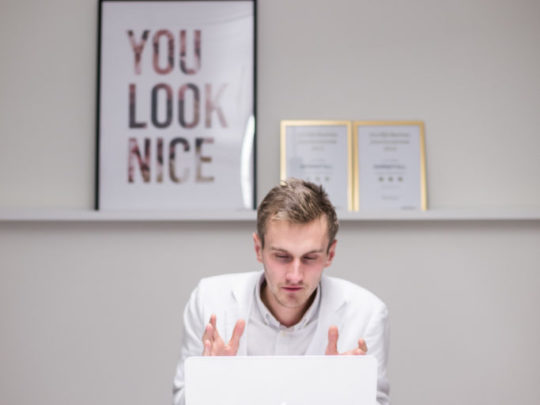 Henning Gillber sitter vid ett konferensbord med sin laptop. Han ser ut att prata med någon genom datorn för han gestikulerar med sina händer.