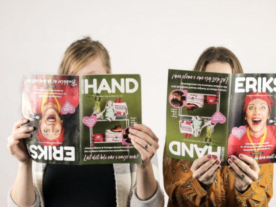 Two people hold the Erikshjälpen newspaper in front of their faces. The magazine can be read from two different angles, so they are holding the magazine at different angles.
