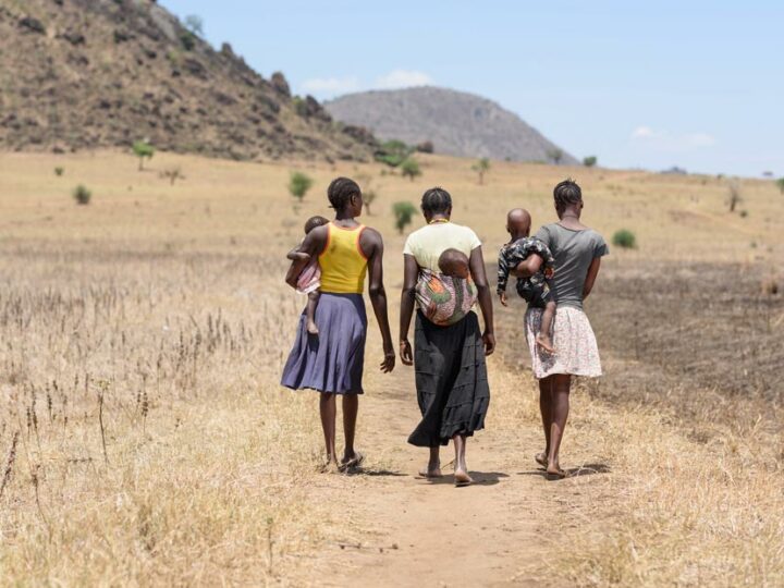 Tre unga kvinnor bärande på varsitt barn vandrar längs en grusväg i Uganda.