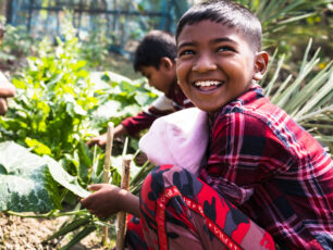4 pojkar sitter i ett grönsaksland i Bangladesh. Killen närmst kameran bär en rutig skjorta. Han tittar upp och ler stort.