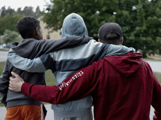 Tre killar bakifrån som håller varandra om ryggen där personen längst ut till höger har en Erikshjälpen-hoodie på sig.