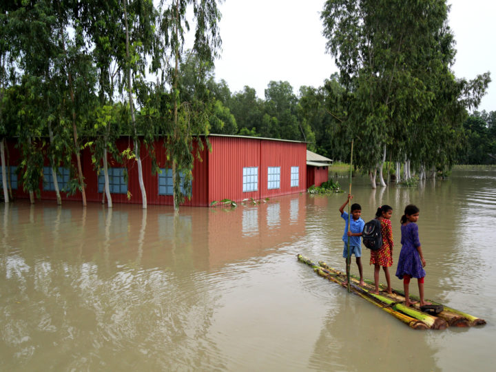 Ett hus står i vatten nästan upp till fönsterna. Tre barn står på en bambuflotte i vattnet. Ett barn ror med en lång pinne. 