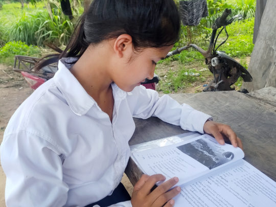 Soeum, en flicka från Kambodja, sitter och läser vid ett bord. Hon syns i profil. I bakgrunden syns grönska i form av buskar och träd.