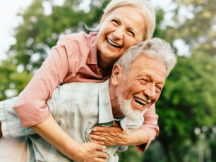 En äldre kvinna rider på en äldre mans rygg. Båda ler.