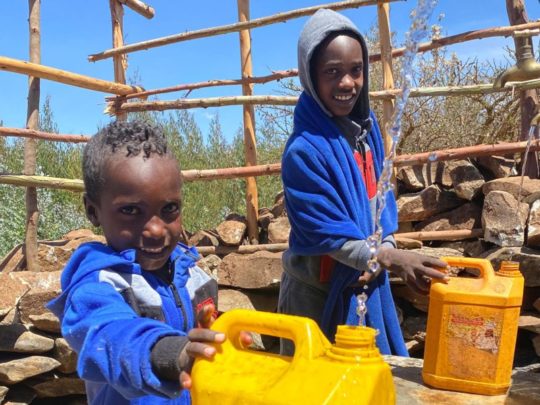Två barn i blåa tröjor står med gula vattenflaskor vid en vattenbrunn i Etiopien