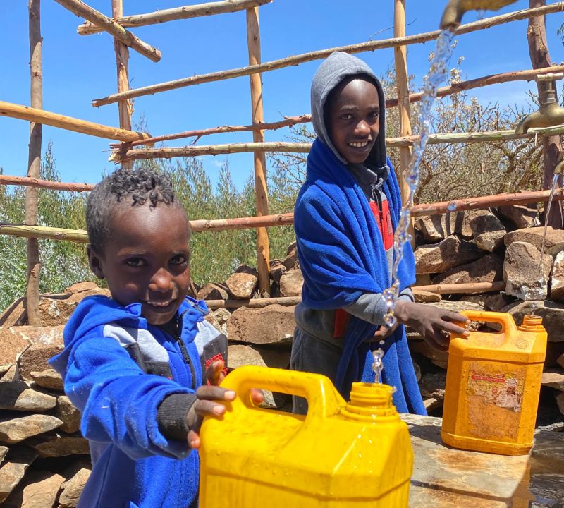 Två barn i blåa tröjor står med gula vattenflaskor vid en vattenbrunn i Etiopien