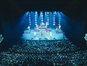 En artist med band står på scen i blått ljus. I publiken sitter mängder med människor.