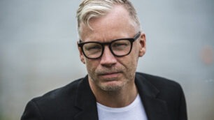 Pressbild på Clas Vårdstedt. Clas har blont hår, svarta glasögon och han är en vit t-short och svart kavaj.