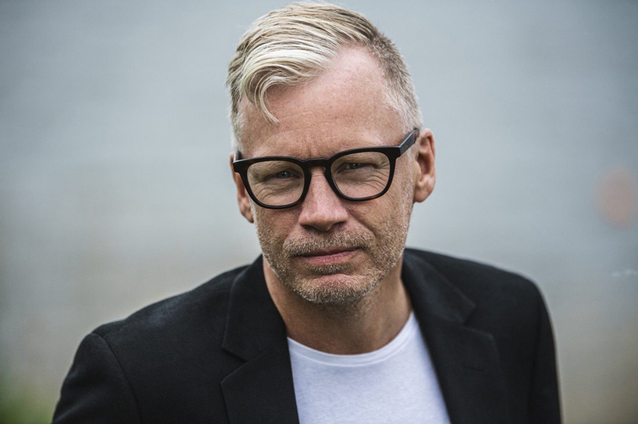 Pressbild på Clas Vårdstedt. Clas har blont hår, svarta glasögon och han är en vit t-short och svart kavaj.