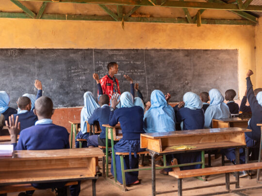 Barn sitter med ryggen mot kameran, vid sina skolbänkar och lyssnar på sin lärare som talar längst fram vid tavlan. Några elever räcker upp händerna.