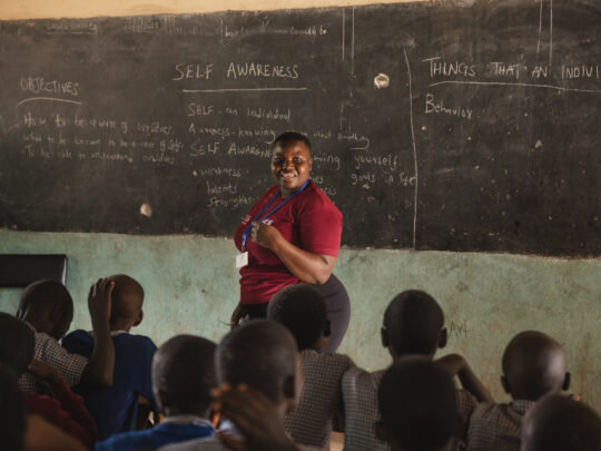 En kvinna står framför en svart griffeltavla i ett klassrum. Framför henne sitter en grupp elever om lyssnar. Kvinnan tittar in i kameran och ler.
