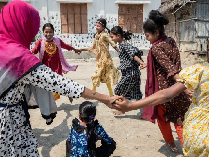 Girls dancing in a circle on a sandy field in Bangladesh.