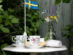 Ett kafebord med två blommiga kaffekoppar, en liten blombukett i en vas och en svenks falgga gjordav trä.