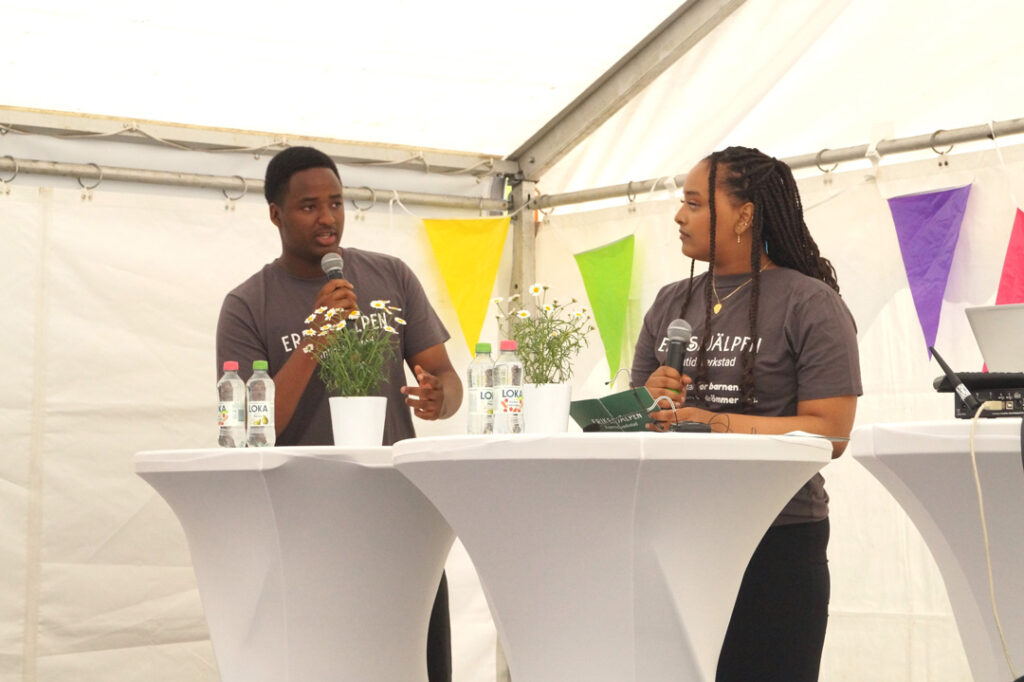 Två ungdomar från Erikshjälpen Framtidsverkstad deltar i ett seminarie under Almedalsveckan. De står vid ett bord på en scen och pratar.