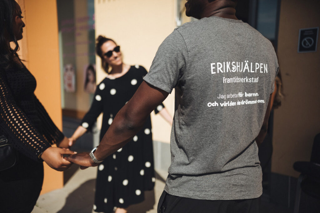 Tre personer står i ring och håller varandra i hand. En av personerna bär en t-shirt som det på ryggen står "Erikshjälpen".