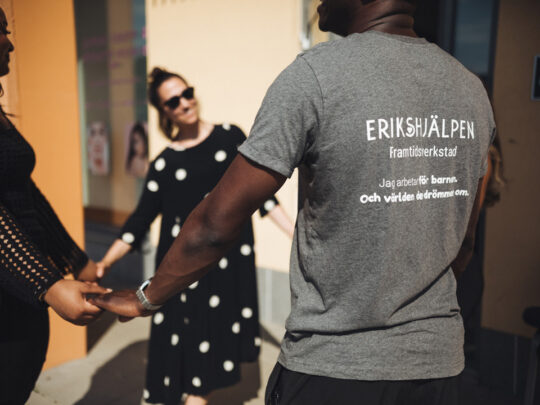 Tre personer står i ring och håller varandra i hand. En av personerna bär en t-shirt som det på ryggen står "Erikshjälpen".