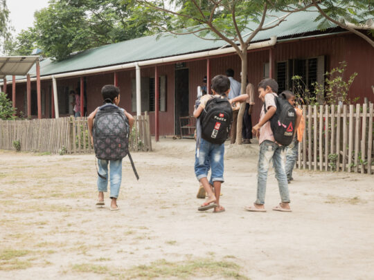Tre killar på en skolgård i Bangladesh. De bär ryggsäckar och är påväg mot klassrummet.