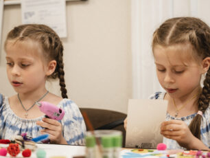 Tvillingsystrarna Alisa och Liya sitter vid ett bord och skapar konst. Alisa håller i en rosa limpistol, medan Liya studerar ett papper. Färgglada pysselmaterial ligger på bordet framför dem.