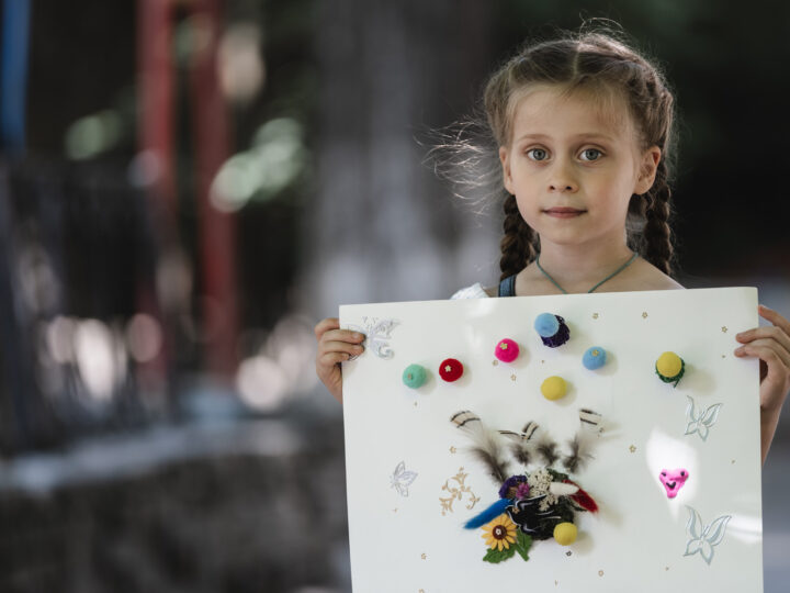 Alisa håller upp ett konstverk dekorerat med färgglada bollar, fjädrar och klistermärken, i en utomhusmiljö med suddig bakgrund.