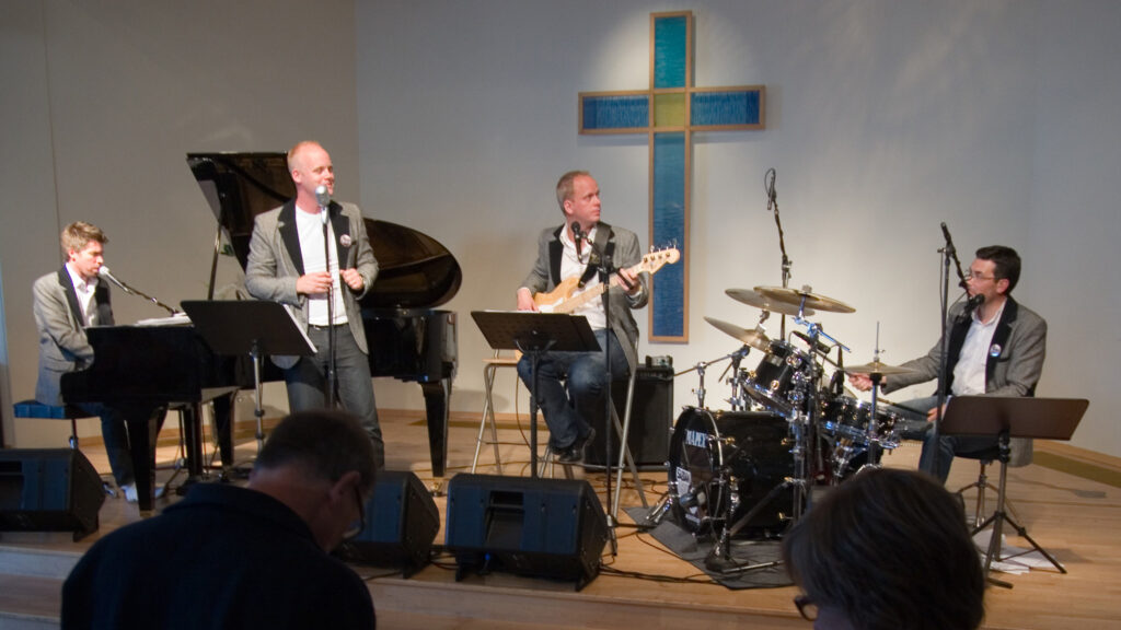 Bandet Memphis Rhythm Chapel uppträder på en scen i en kyrka. De är fyra i bandet. En spelar piano, en sjunger, en spelar gitarr och en spelar trummor. Bandet bär matchande grå kavajer.