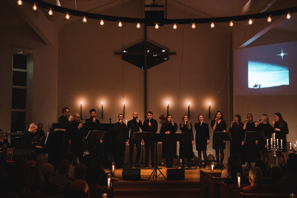 En kör står på rad i en kyrka och sjunger till publiken. Det är levande ljus tända och alla personer i kören bär svart.