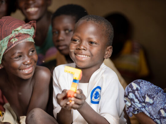 Ali, en 6-årig pojke sitter med en leksak i handen. Han ler stort. Bredvid honom sitter hans kompis som tittar på Ali och ler.