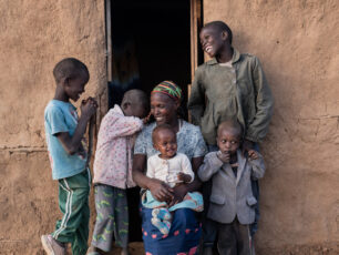 En familj med 5 barn och deras mamma står framför ingången till deras hem, en hydda i Afrika. Familjen ler.