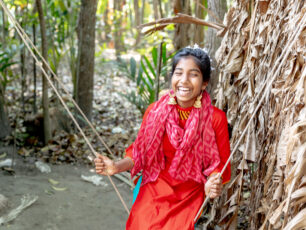 En flicka i Bangladesh gungar. Hon bär en röd klännin goch skrattar. I bakgrunden syns natur.