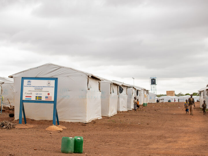 Ett flyktingläger i Burkina Faso. Tält gjorda av presseningar står uppradade. Marken består av saknd oh grus. Himlen är grå och dyster. Några människor syns komma gående mellan tälten.