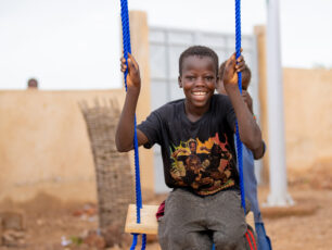 Ali 11 år gungar på en gunga på en grusplan i Burkina Faso. Han tittar in i kameran och ler stort.