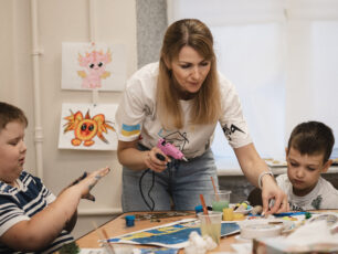 Barnpsykologen Lyudmila Burtseva i en vit tröja och jeans, som hjälper två barn att limma under konstterapi.