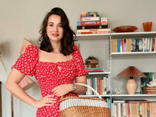 Sara Olsson in a red dress, carrying a wicker basket in front of a bookshelf.