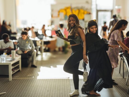 Two young women dance in a room full of young people.