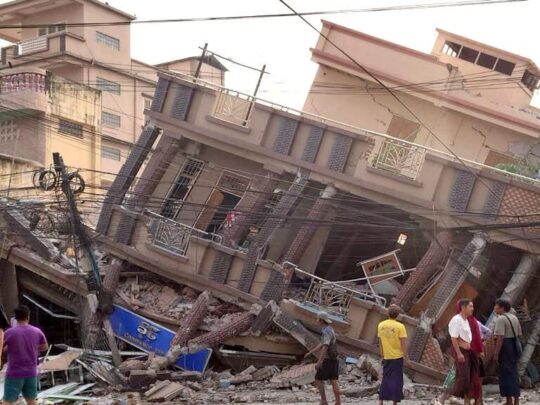 People standing in ruins in front of a house partially toppled by the earthquake.