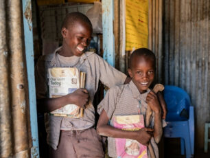 Två barn står i en byggnad i Kenya med skolböcker i famnen och ler.