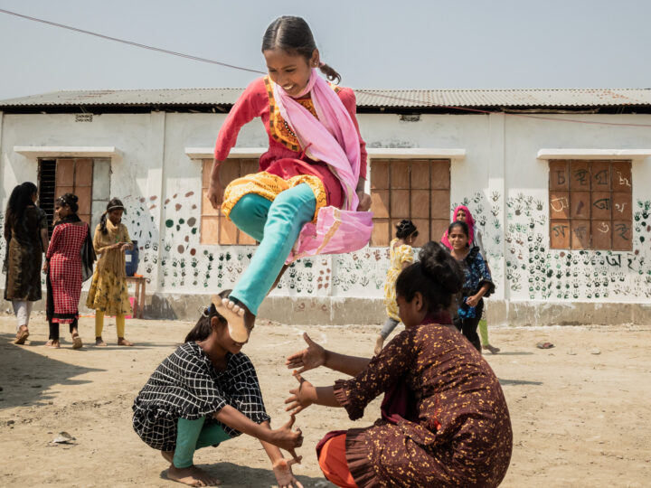 En flicka hoppar glädjefullt över sina vänners händer på en torr gårdsplan i Bangladesh.