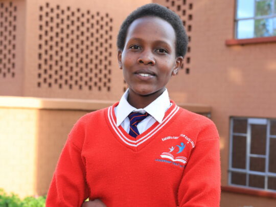 Nancy Mbiti som står i sin skoluniform utanför en tegelbyggnad.