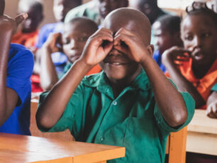 En pojke i grön skjorta som sitter i en skolbänk och formar händerna som en kikare framför ögonen.