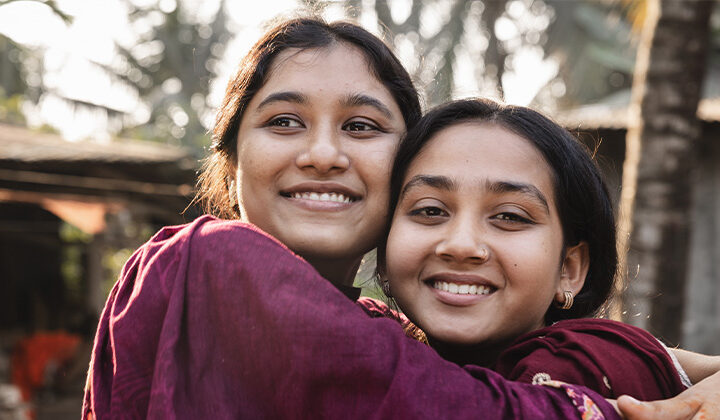 Två flickor kramas och ler mot kameran i solig utomhusmiljö i Bangladesh.