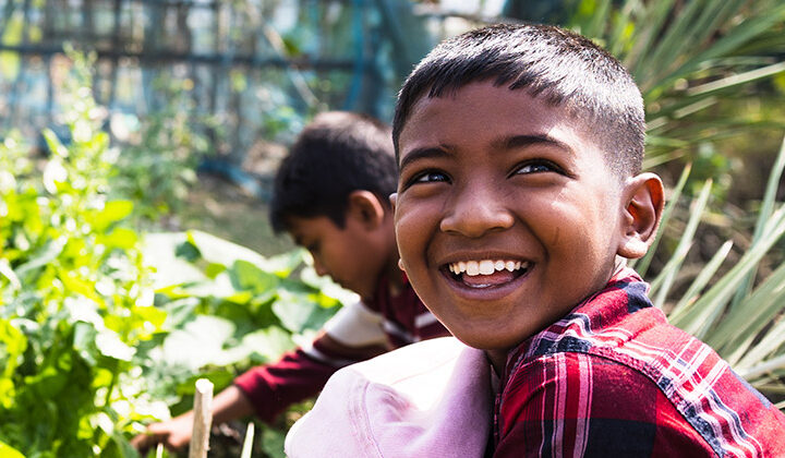Leende pojke i röd- och svart-rutig skjorta sitter på huk i en trädgård, med en annan pojke i bakgrunden som plockar grönsaker.