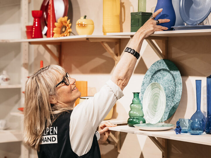 Butiksbiträde plockar ner en blå vas från en hylla fylld med färgglad inredning.