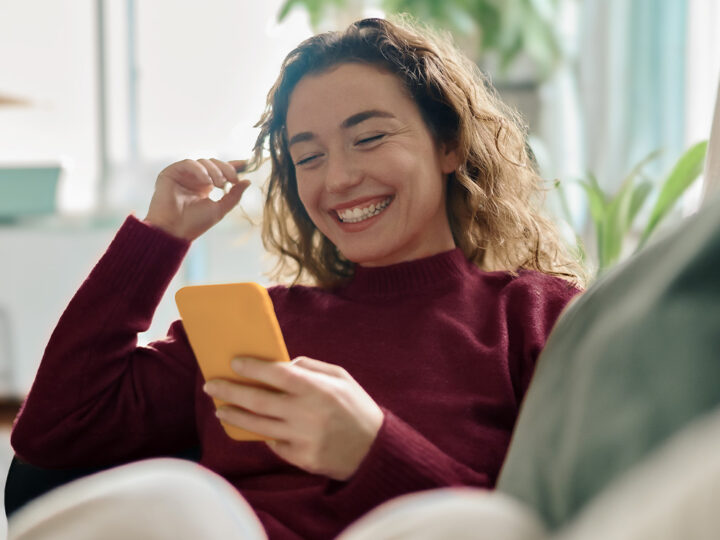 Ung kvinna i vinröd stickad tröja sitter i en soffa i ett ljust rum och ler stort medan hon tittar på sin smartphone.
