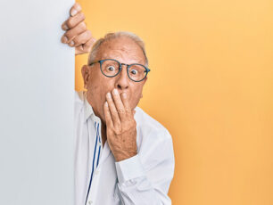 En äldre man kikar fram bakom en vit vägg framför en gul bakgrund och håller sig för munnen med ett lekfullt, förvånat uttryck.