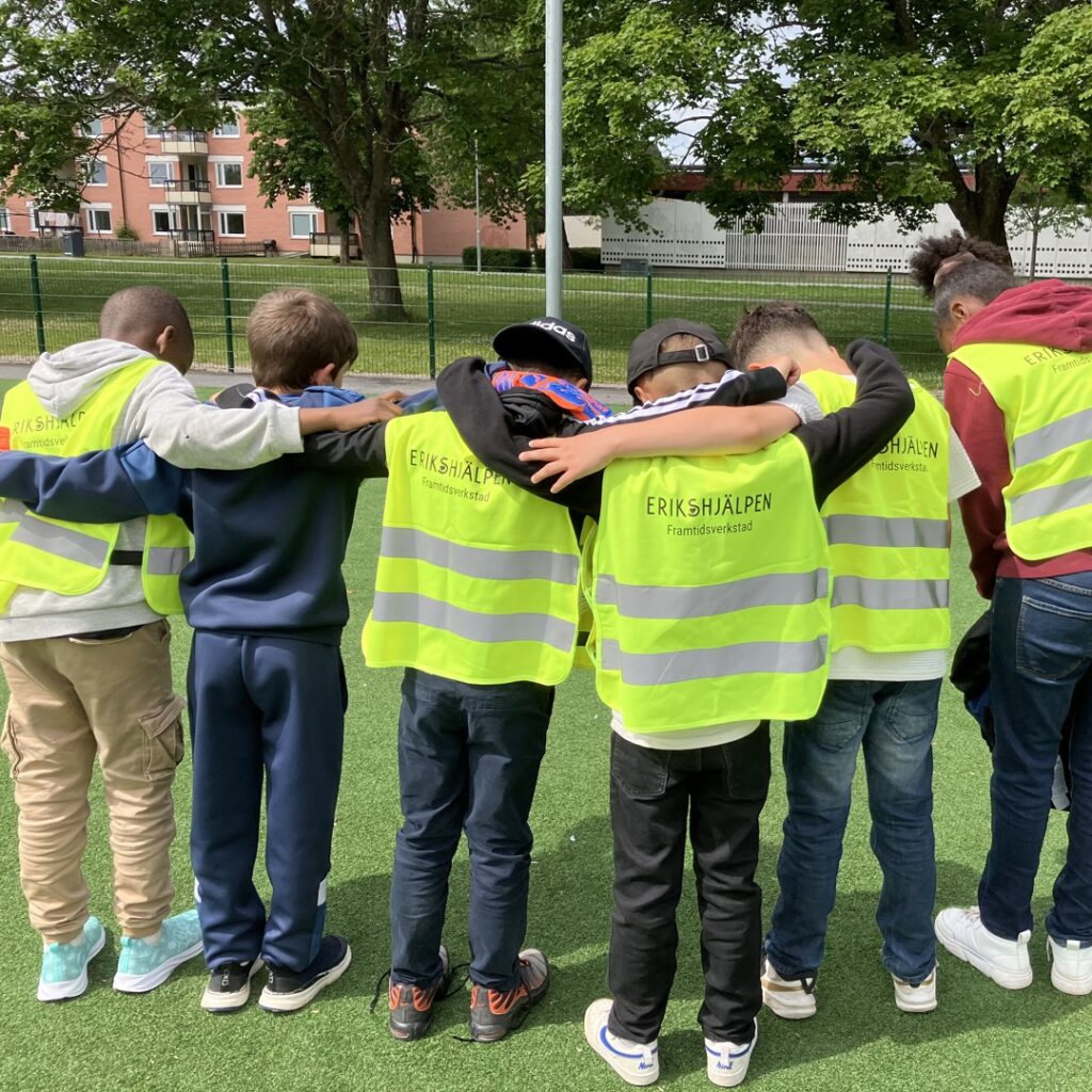 Barn som bär reflexvästar med texten Erikshjälpen Framtidsverkstad på ryggen står på led och håller varandra om axlarna.