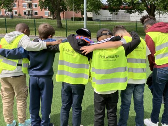 Barn som bär reflexvästar med texten Erikshjälpen Framtidsverkstad på ryggen står på led och håller varandra om axlarna.