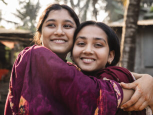 Två flickor kramas och ler mot kameran i solig utomhusmiljö i Bangladesh.
