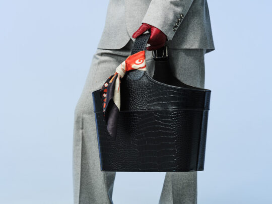 Person i grå kostym som håller en svart väska. En färgglad scarf är knuten runt handtaget och personen bär röda handskar.