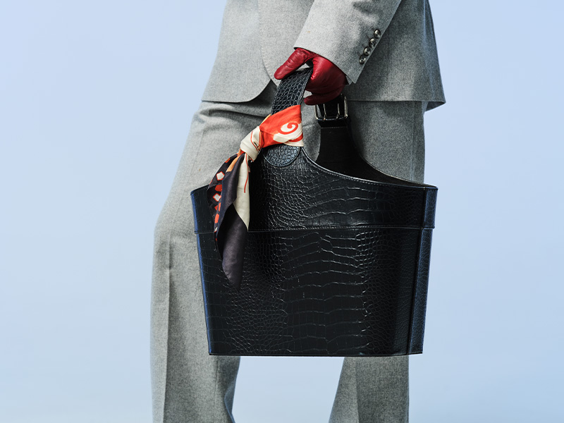 Person i grå kostym som håller en svart väska. En färgglad scarf är knuten runt handtaget och personen bär röda handskar.
