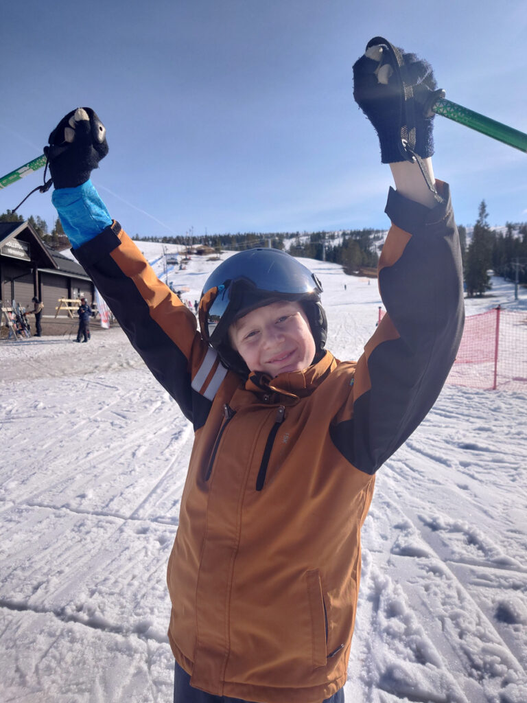 Svante står i en solig skidbacke och lyfter händerna i en segergest och ler mot kameran.
