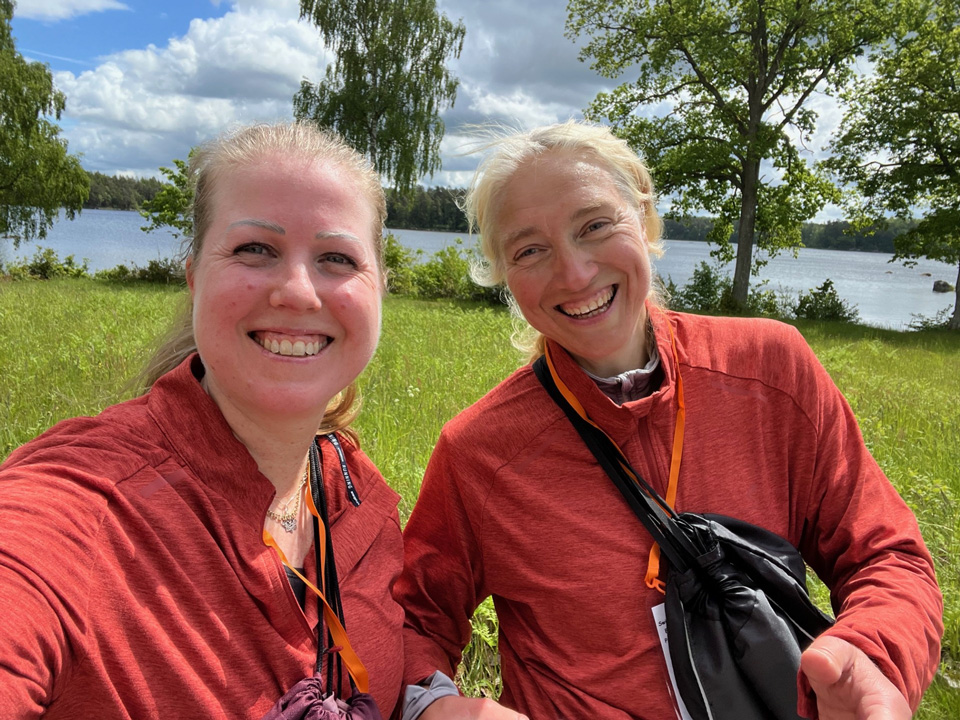 Therese Blåsol och Helena Simonsson ler mot kameran i solig utomhusmiljö. Grönskande träd och en sjö syns i bakgrunden.